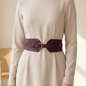 May include: A long-sleeved, floor-length beige dress featuring a round neckline and a fitted waist. The dress is cinched with a wide, dark burgundy corset-style belt that has a central silver-toned buckle and a structured, overlapping design.