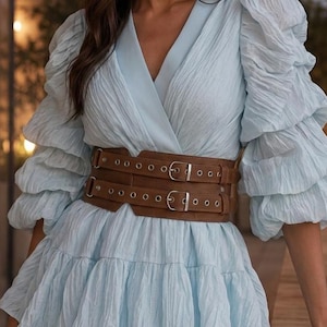 May include: A light blue long-sleeved dress with a textured, crinkled fabric, a V-neckline, and tiered skirt sections. The dress is cinched at the waist with a wide, brown, double-buckle belt featuring silver-toned metal hardware and eyelets.