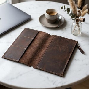 Peut inclure: Un portefeuille ouvert en cuir marron foncé avec une surface texturée, des coutures visibles sur les bords, trois fentes pour cartes sur le côté gauche et un compartiment zippé sur le côté droit, posé à plat sur une surface.
