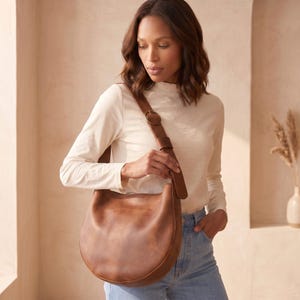 K&ouml;nnte beinhalten: Eine braune Ledertasche mit geschwungenem Design. Die Tasche hat einen langen Riemen mit einer kreisf&ouml;rmigen Schnalle. Die Tasche wird &uuml;ber der Schulter getragen. Das Model tr&auml;gt ein wei&szlig;es Langarmshirt und blaue Jeans.