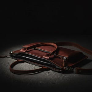 May include: A brown leather laptop bag with a top handle and a detachable shoulder strap. The bag has a zipper closure and a sleek, minimalist design. The bag is open, revealing a laptop inside.
