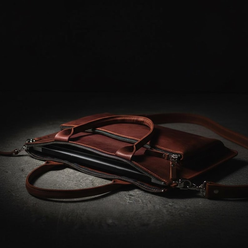 May include: A brown leather laptop bag with a top handle and a detachable shoulder strap. The bag has a zipper closure and a sleek, minimalist design. The bag is open, revealing a laptop inside.