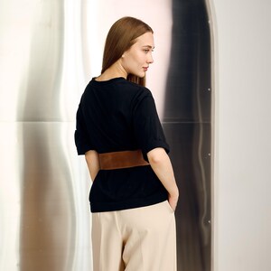May include: A black t-shirt with rolled-up sleeves and a wide brown belt. The shirt is paired with light beige pants. The background is a shiny, metallic surface.