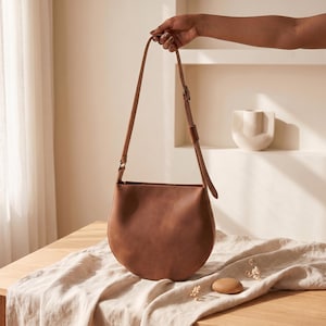 K&ouml;nnte beinhalten: Eine braune Leder-Umh&auml;ngetasche mit gebogenem Boden und einem langen, verstellbaren Riemen. Die Tasche wird von einer Hand gehalten. Die Tasche liegt auf einem Holztisch mit einem beigen Tuch.