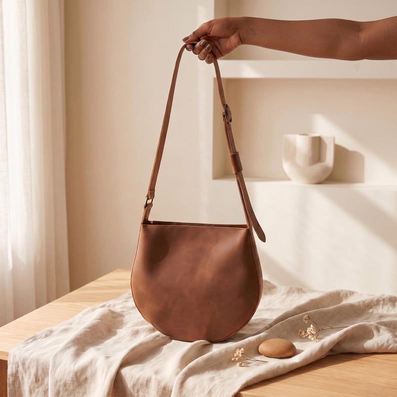 K&ouml;nnte beinhalten: Eine braune Leder-Umh&auml;ngetasche mit gebogenem Boden und einem langen, verstellbaren Riemen. Die Tasche wird von einer Hand gehalten. Die Tasche liegt auf einem Holztisch mit einem beigen Tuch.