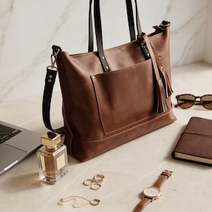 May include: A brown leather tote bag with black handles and a detachable shoulder strap. The bag has a front pocket and a decorative tassel. Accessories include sunglasses, a notebook, a watch, jewelry, and a perfume bottle.