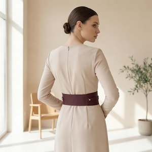 May include: A long-sleeved, beige midi dress featuring a round neckline and a fitted silhouette. The dress is cinched at the waist with a wide, dark burgundy belt that includes a horizontal strap detail and a small silver-toned metal stud closure.