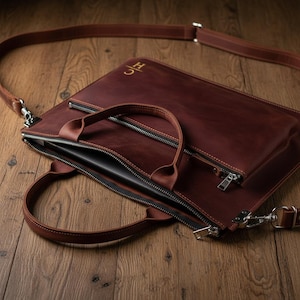 May include: A brown leather briefcase with a top handle and a detachable shoulder strap. The briefcase has a zippered main compartment and a smaller zippered pocket on the front. The initials "JC" are embossed in gold on the upper right corner.