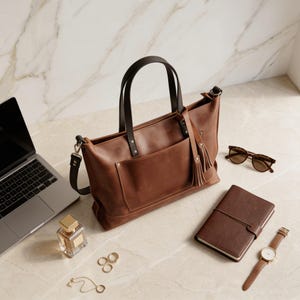 May include: A brown leather tote bag with a front pocket and a tassel, next to a laptop, sunglasses, a notebook, a watch, perfume, and jewelry. The bag has a dark brown handle and a shoulder strap.