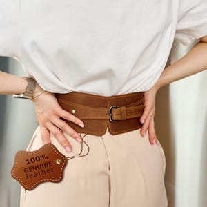 Op de afbeelding: Een brede bruine leren riem met een zilveren gesp. De riem wordt om de taille gedragen. Een label met de tekst "100% ECHT LEER" is aan de riem bevestigd.