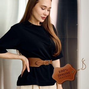 May include: A wide, brown leather belt with a silver buckle. The belt is worn around the waist. A tag with the text "100% GENUINE leather" is attached to the belt.