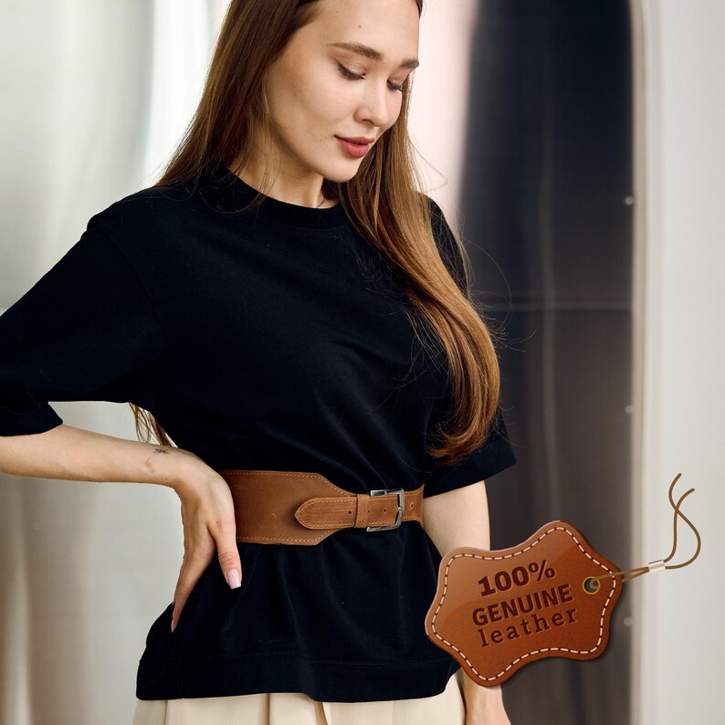 May include: A wide, brown leather belt with a silver buckle. The belt is worn around the waist. A tag with the text "100% GENUINE leather" is attached to the belt.