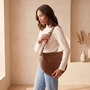 K&ouml;nnte beinhalten: Eine braune Leder-Umh&auml;ngetasche mit Halbmondform. Die Tasche hat einen langen Riemen und eine runde Schnalle. Die Frau auf dem Bild tr&auml;gt ein wei&szlig;es Langarmshirt und blaue Jeans.