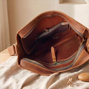 K&ouml;nnte beinhalten: Ge&ouml;ffnete braune Leder-Umh&auml;ngetasche mit mehreren Innentaschen und einem Rei&szlig;verschluss. Die Tasche hat einen langen, verstellbaren Riemen und ein abgenutztes Aussehen. Das Innere ist in einem helleren Braunton gehalten.