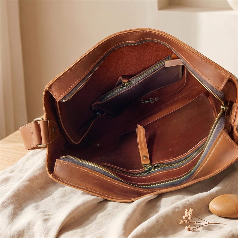 K&ouml;nnte beinhalten: Ge&ouml;ffnete braune Leder-Umh&auml;ngetasche mit mehreren Innentaschen und einem Rei&szlig;verschluss. Die Tasche hat einen langen, verstellbaren Riemen und ein abgenutztes Aussehen. Das Innere ist in einem helleren Braunton gehalten.
