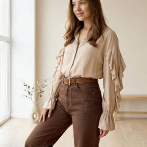 Pu&ograve; includere: Una camicia beige abbottonata con maniche a balze e una cintura marrone, abbinata a pantaloni marroni. La camicia ha un colletto classico e polsini abbottonati. La cintura ha una fibbia argentata. I pantaloni sono a vita alta.