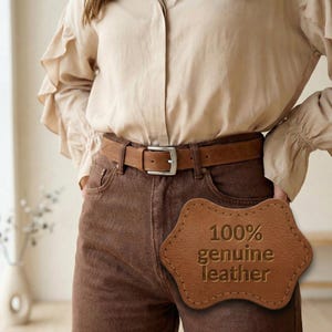 Pu&ograve; includere: Una cintura in pelle marrone con fibbia argentata &egrave; indossata con pantaloni marroni e una camicia beige abbottonata. Un'etichetta in pelle con la scritta "100% genuine leather" &egrave; visibile. La cintura &egrave; un accessorio classico.