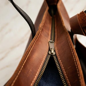 May include: Close-up of a brown leather tote bag interior with a brass zipper. The bag has a black strap and a textured, distressed appearance. The interior appears to be lined with a dark blue fabric. The stitching is visible along the edges.