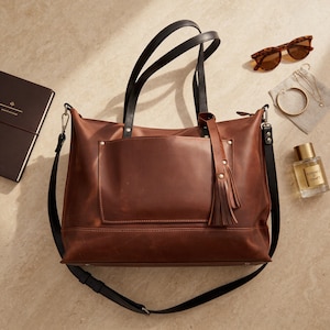 May include: A brown leather tote bag with black handles and a detachable shoulder strap. The bag has a front pocket and a tassel. Accessories include sunglasses, a notebook, jewelry, and a perfume bottle.
