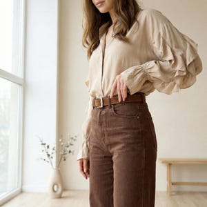 Pu&ograve; includere: Una camicia beige abbottonata con maniche a balze e pantaloni di velluto a coste marroni. Una cintura in pelle marrone con fibbia argentata &egrave; indossata con i pantaloni. L'outfit &egrave; in una palette di colori neutri.
