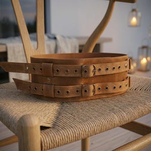 May include: A wide tan leather corset belt featuring two horizontal straps with brass-toned metal buckles and matching grommets. The belt has a smooth, matte finish and is constructed with two distinct layers, each secured by its own buckle.
