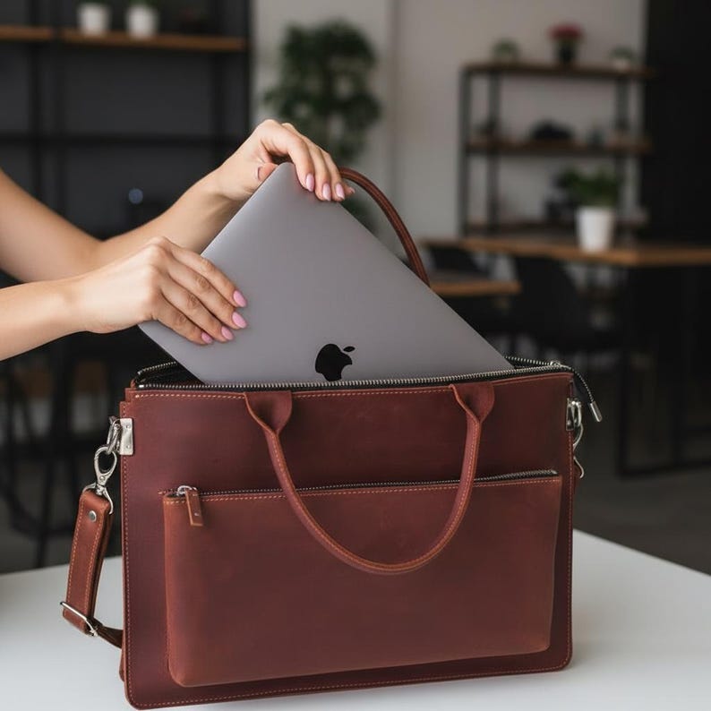 May include: A brown leather laptop bag with a silver zipper and a front pocket. A gray laptop with the Apple logo is being placed inside the bag. The bag has a handle and a shoulder strap. The background is a blurred office setting.