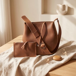 K&ouml;nnte beinhalten: Eine braune Leder-Umh&auml;ngetasche und eine passende kleine Tasche. Die Tasche hat einen breiten, verstellbaren Riemen und eine gro&szlig;e silberne Schnalle. Die Tasche hat einen kleinen Riemen mit einem Knoten. Das Set liegt auf einem beigen Leinentuch.