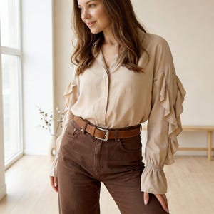 Pu&ograve; includere: Una camicia beige abbottonata con maniche a balze e una cintura marrone. La camicia &egrave; abbinata a pantaloni marroni. L'outfit &egrave; semplice ed elegante, adatto a occasioni casual o semi-formali.