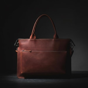 May include: A brown leather briefcase with a top handle and a front zippered pocket. The bag has a rectangular shape and a shoulder strap attachment. The leather has a smooth texture and a rich, warm color.