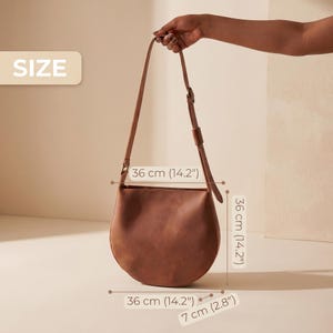 K&ouml;nnte beinhalten: Eine braune Leder-Umh&auml;ngetasche mit abgerundetem Boden und verstellbarem Riemen. Die Tasche ist 36 cm breit, 36 cm hoch und 7 cm tief. Die Tasche wird von einer Hand gehalten.