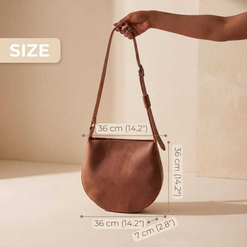 K&ouml;nnte beinhalten: Eine braune Leder-Umh&auml;ngetasche mit abgerundetem Boden und verstellbarem Riemen. Die Tasche ist 36 cm breit, 36 cm hoch und 7 cm tief. Die Tasche wird von einer Hand gehalten.