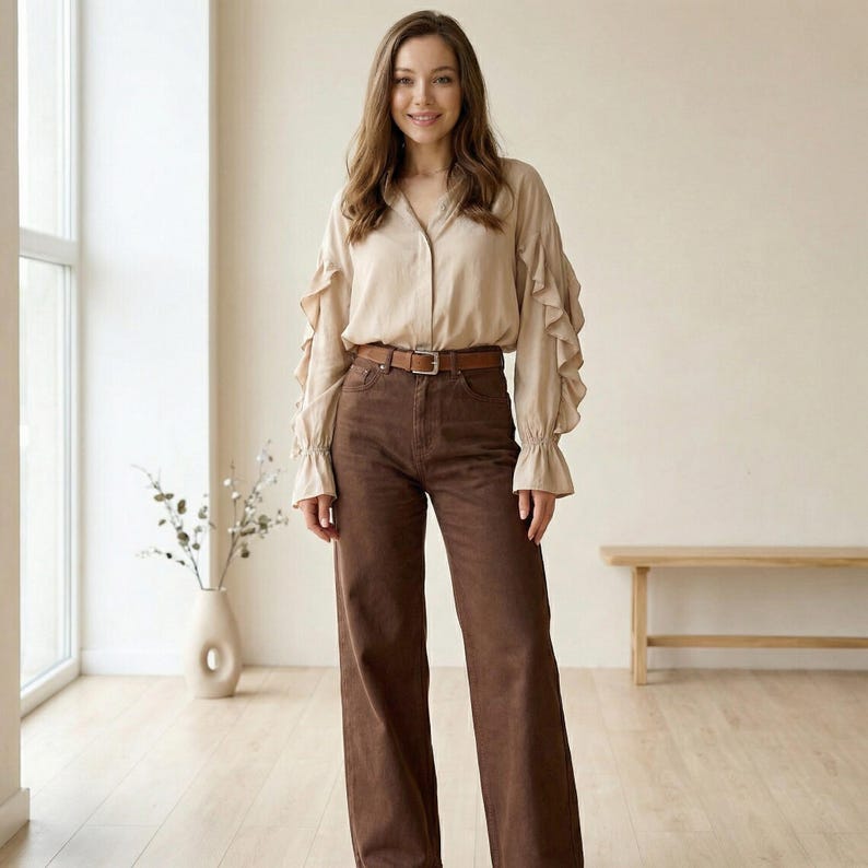 Pu&ograve; includere: Una donna indossa una camicetta beige con maniche a balze e pantaloni marroni a gamba larga. L'outfit &egrave; impreziosito da una cintura marrone. Lo sfondo comprende una finestra, un vaso con rami e una panca in legno.