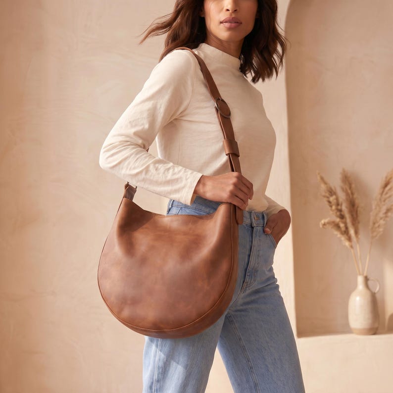 K&ouml;nnte beinhalten: Eine braune Leder-Umh&auml;ngetasche mit geschwungener Form. Die Tasche hat einen verstellbaren Riemen und eine Schnalle. Die Person tr&auml;gt ein wei&szlig;es Langarmshirt und blaue Jeans.