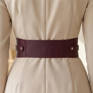 May include: A wide, dark burgundy waist belt worn over a beige blazer. The belt features a smooth, solid-colored surface with two horizontal side straps, each secured by a single silver-toned metal stud, creating a structured and clean appearance.