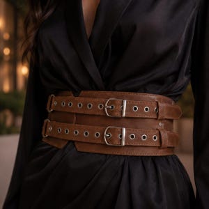 May include: A wide brown belt featuring two horizontal straps with silver-toned metal buckles and rows of grommets. The belt is constructed from a smooth, matte material and is designed to wrap around the waist, providing a structured appearance.