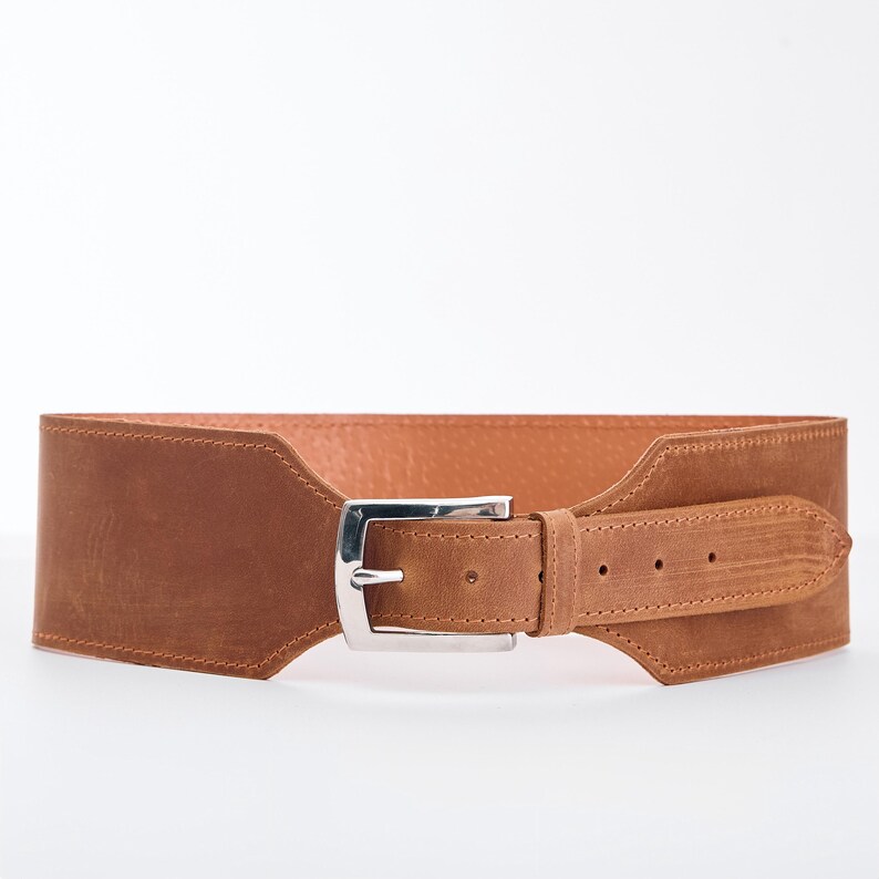 May include: A wide, brown leather belt with a silver-colored buckle. The belt has a suede-like texture and a rectangular shape. The buckle is a classic design, and the belt appears to be new.