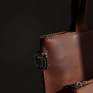 May include: Close-up of a brown leather tote bag with a silver-colored metal clasp and zipper. The bag has a handle and a zipper closure. The leather has visible stitching.