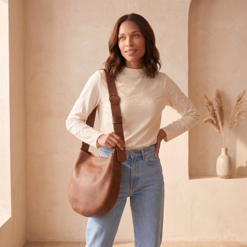 K&ouml;nnte beinhalten: Eine braune Schultertasche aus Leder mit geschwungenem Design. Die Tasche hat einen breiten Riemen mit einer kreisf&ouml;rmigen Schnalle. Die Tasche wird &uuml;ber der Schulter getragen. Das Model tr&auml;gt ein cremefarbenes Langarmshirt und blaue Jeans.