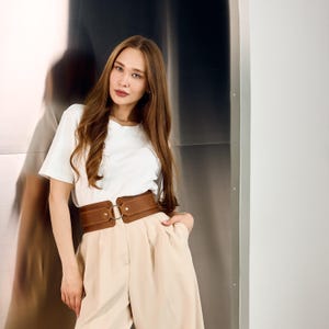 Op de afbeelding: Een vrouw draagt een wit T-shirt en een beige broek, met een brede bruine leren riem die de taille accentueert. De riem heeft een grote, zilverkleurige ringgesp. De achtergrond is een metalen muur.