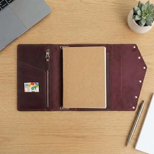 Puede incluir: Una funda de cuero color burdeos oscuro para cuaderno con un bolsillo vertical con cremallera a la izquierda, una banda elástica central que sujeta un cuaderno de papel beige y un cierre de solapa puntiaguda con remaches metálicos.