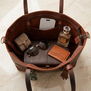 May include: A brown leather tote bag filled with accessories. Inside are sunglasses, a smartphone, a small watch case, a leather wallet, a journal, and a perfume bottle. The bag has a zipper closure and two handles.