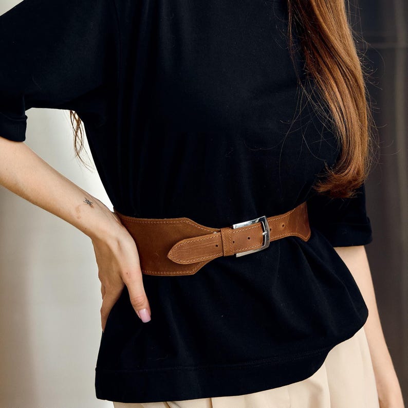 May include: A wide, brown leather belt with a silver-colored buckle. The belt is worn over a black top and beige pants. The belt has a wide, curved design at the front.