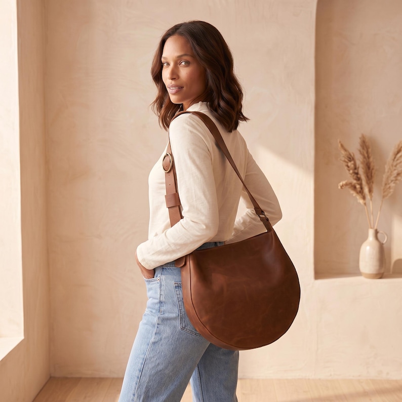 K&ouml;nnte beinhalten: Eine braune Ledertasche mit einem geschwungenen Design. Die Tasche hat einen langen Riemen und einen kleineren Riemen mit einer Schnalle. Die Person tr&auml;gt ein wei&szlig;es Langarmshirt und blaue Jeans.