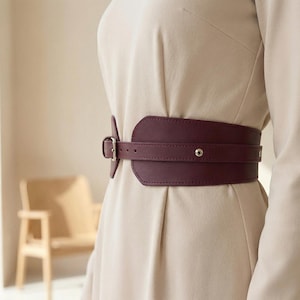 May include: A wide, deep burgundy waist belt featuring a smooth finish, a central horizontal strap, and a silver-toned metal buckle closure. The belt is worn over a plain, long-sleeved beige dress, highlighting its structured, contoured shape.