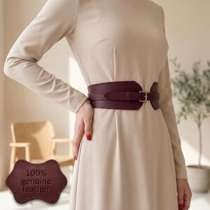 May include: A wide, dark burgundy leather waist belt featuring a silver-toned metal buckle and a matching strap loop. A separate, scalloped-edge leather tag is visible in the corner, embossed with the text 100% genuine leather.