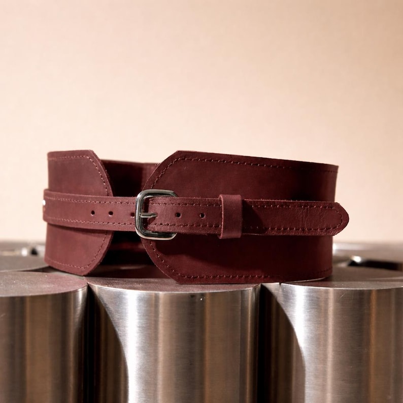 May include: A wide, dark maroon leather corset belt featuring a smooth matte finish, a central horizontal strap with visible stitching, and a polished silver-tone metal buckle with multiple adjustment holes for a secure fit around the waist.