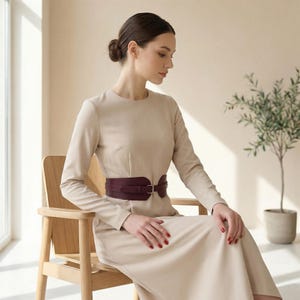 May include: A long-sleeved beige midi dress featuring a round neckline and a wide, dark burgundy waist belt with a silver-toned metal buckle. The dress has a fitted bodice and a flowing skirt, creating a structured yet elegant silhouette.