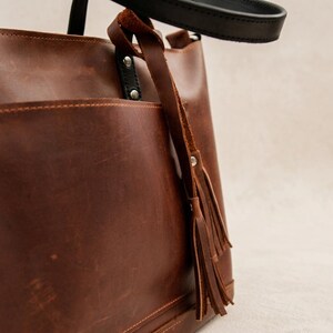 May include: A brown leather tote bag with a black handle and a decorative tassel. The bag has a large pocket on the front and is made of a rich, textured leather. The bag is designed for everyday use.