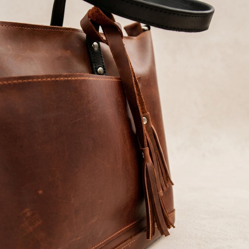 May include: A brown leather tote bag with a black handle and a decorative tassel. The bag has a large pocket on the front and is made of a rich, textured leather. The bag is designed for everyday use.