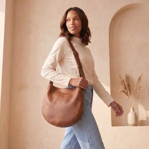 K&ouml;nnte beinhalten: Eine braune Schultertasche aus Leder mit geschwungenem Design. Die Tasche hat einen langen Riemen und eine runde Schnalle. Die Tasche wird &uuml;ber der Schulter getragen. Der Hintergrund ist neutral.